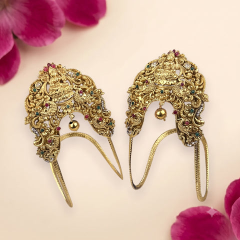 A pair of ornate gold-toned traditional Indian hair ornaments (jhumkas) decorated with intricate filigree, small hanging bells, and colorful rhinestones, displayed on a light pink background with pink flower petals in the corners.