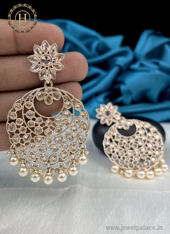 A pair of gold-plated kundan earrings with pearl studs, held in a hand, against a blue fabric backdrop.